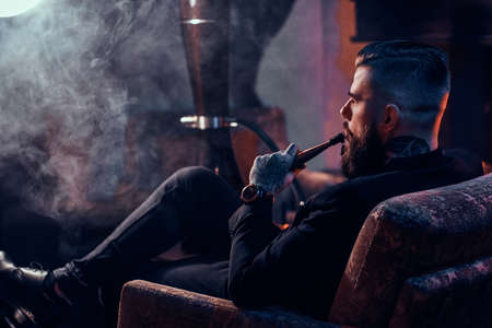 Attractive bearded man is making nice misty vapour while relaxing near fireplace and smoking hookah.の写真素材