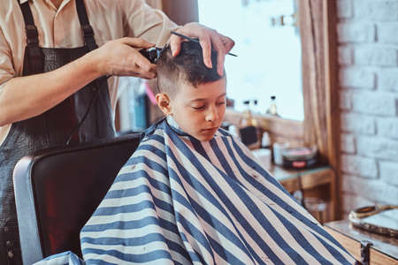 Young school boy is getting trendy hairstyle from expirienced hairdresser at modern barbershop.