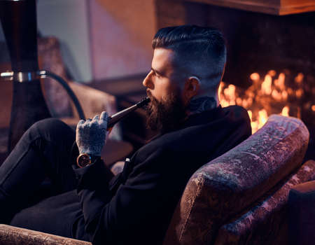 Groomed bearded man is relaxing on lounge near fireplace while smoking hookah. He has tattoo on his hand.の写真素材