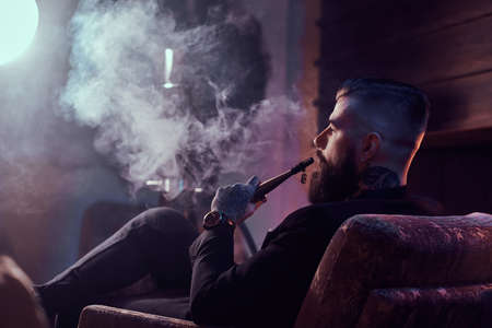 Attractive bearded man is making nice misty vapour while relaxing near fireplace and smoking hookah.の写真素材