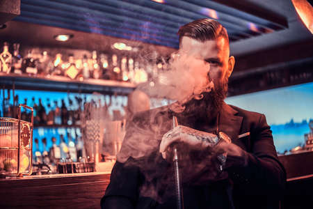 Portrait of tattooed bearded man which is smoking hookah, making nice vapour while sitting near bar...