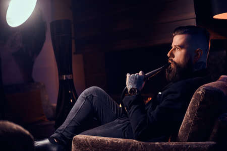 Groomed tattooed man is relaxing near fireplace in dark room and smoking hookah.