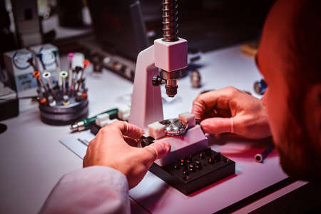 Mature watchmaker is doing engraving for custmers watch at his workshop.