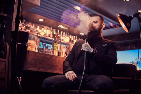 Portrait of tattooed bearded man which is smoking hookah, making nice vapour while sitting near bar...