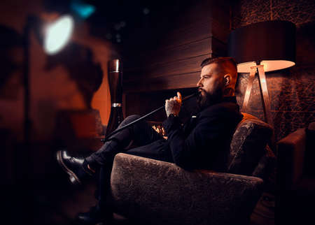 Handsome bearded man is relaxing on lounge while smoking hookah. He has tattoo on his hand.
