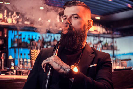 Portrait of tattooed bearded man which is smoking hookah, making nice vapour while sitting near bar...
