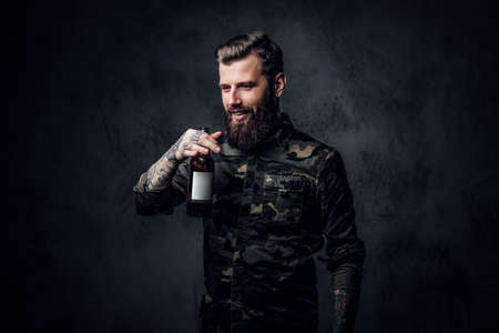 Stylish bearded hipster guy in military shirt enjoys the taste of craft beer. Studio photo against a dark wallの写真素材