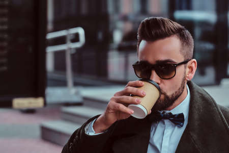 Attractive elegant man in sunglasses is drinking his coffee while sitting outside at coffeeshop.の写真素材