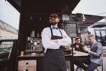 Groomed elegant barista in sunglasses is standing crossed his hands next to his small coffeeshop.の写真素材