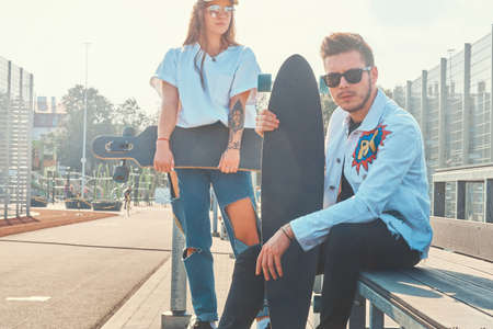Young trendy couple is chilling on the sunny street with their longboards. They wear denim.