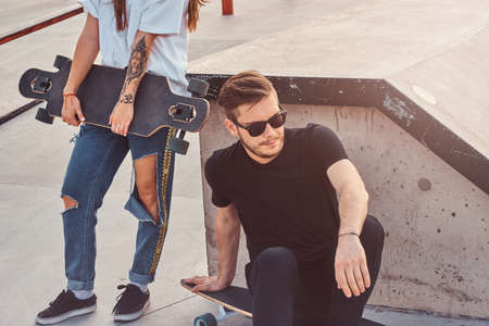 Young trendy couple is sitting at sunny skatepark with their longboards. There is ramp at background.の写真素材