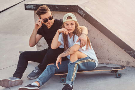 Young trendy couple is sitting at sunny skatepark with their longboards. There is ramp at background.の写真素材