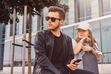 Young attractive couple is sitting on the summer street with their longboard. Man is showing...