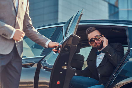 Attractive groomed man in sunglasses is talking by smartphone and sitting in the car while his assistant is opening door for him.の写真素材