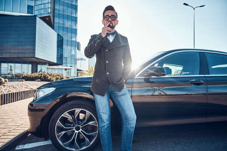 Handsome man in coat is standing near his new car while smoking at the parking.