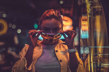Beautiful pensive trendy girl is posing for photographer in neon lights. There are darkness at background.の写真素材