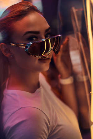 Portrait of pensive trendy woman in sunglasses and white tshirt with pink neon lights.