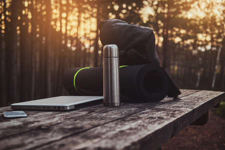 Silver laptop, smartphone , black backpack, mat and thermos on the wooden table in the forest. Sunset at background.の写真素材