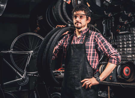 Pensive happy man in protective glasses is standing with tires at bicycle repairing studio.の写真素材