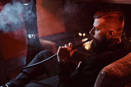 Groomed bearded man is relaxing on lounge near fireplace while smoking hookah. He has tattoo on his hand.の写真素材
