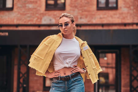 Attractive young woman in sunglasses and yellow jacket is posing for photographer at random city yard.の写真素材