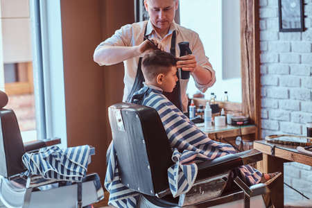 Young school boy is getting trendy hairstyle from expirienced hairdresser at modern barbershop.の写真素材
