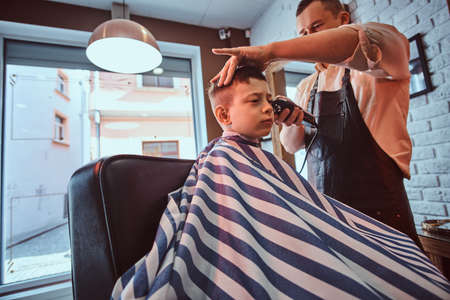 Cute little child is getting trendy haircut from barber at busy barbershop.の写真素材