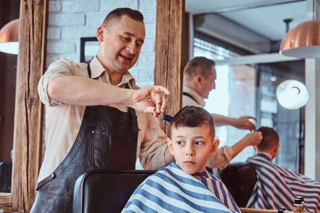 Attractive school boy is getting trendy haircut from mature hairdresser at fashionable hairdressing salon.の写真素材