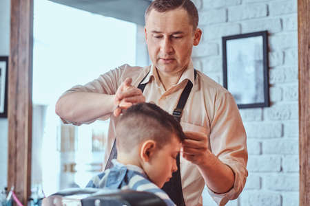 Attractive school boy is getting trendy haircut from mature hairdresser at fashionable hairdressing salon.の写真素材