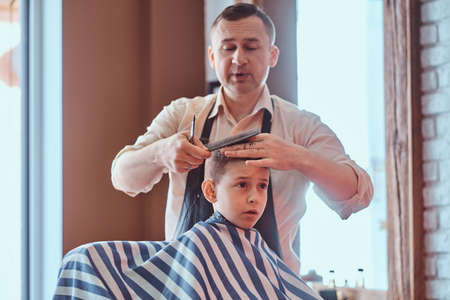 Small charming boy is getting trendy haircut from expirienced barber at fashionable hairdressing salon.の写真素材