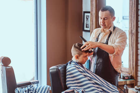 Young school boy is getting trendy hairstyle from expirienced hairdresser at modern barbershop.