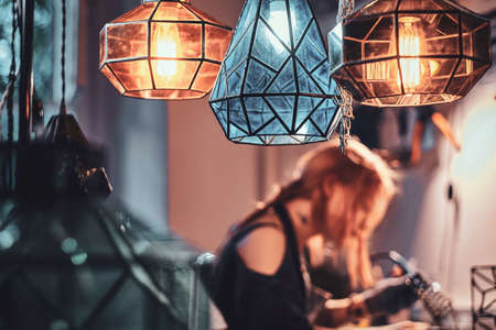 There are beautiful handmade lamps from colourful glass at workshop and Woman is working with soldering iron at background.の写真素材