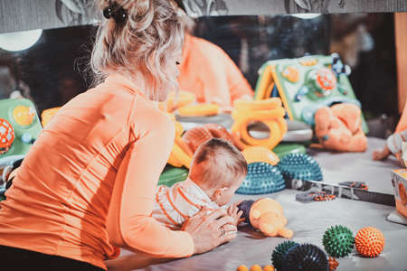 Young happy mom is plaing with her newborn baby at masseur cabinet full of special treatment toys.