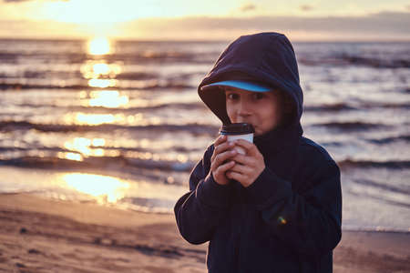 Pretty little boy in cap and hood is drinking tea at seaside. There are beautiful sunset at background.の写真素材