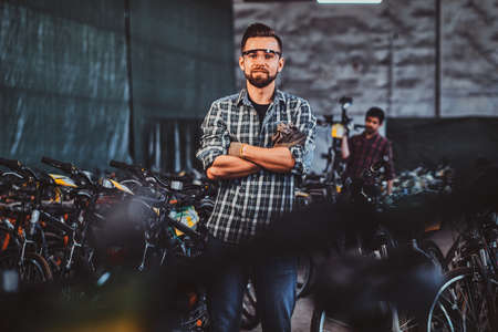 Pensive man in protective glasses and checkered shirt is posing at his own warehouse full of bicycles.の写真素材