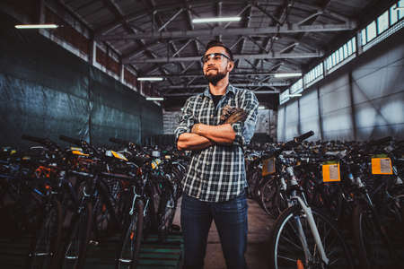 Pensive man in protective glasses and checkered shirt is posing at his own warehouse full of bicycles.の写真素材