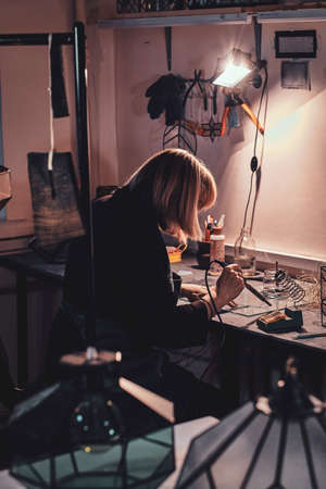 Expirienced designer is working with soldering iron at her own cozy workshop.