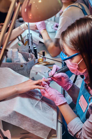 Focused talented manicurist is working at her own workplace at busy beauty salon. She is wearing protective mask.の写真素材