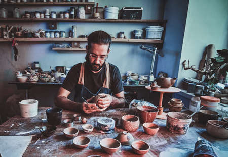 Pensive entusiastici potter in glasses at his own workshop is workig on a new handmade teapot.の写真素材