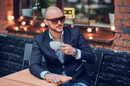 Pensive groomed man is enjoying coffee while sitting outside at cafe on his own break.の写真素材