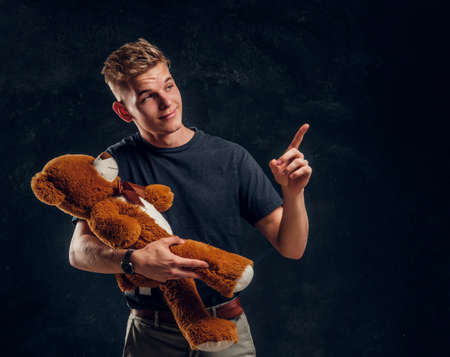 Young attractive man is showing something exiting for his friend giand toy bear at dark studio.の写真素材