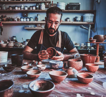 Pensive entusiastici potter in glasses at his own workshop is workig on a new handmade teapot.の写真素材