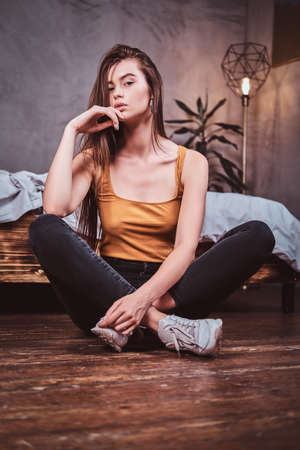 Beautiful sexy girl is sitting on the floor at her bedroom. She has casual style and long hair.の写真素材
