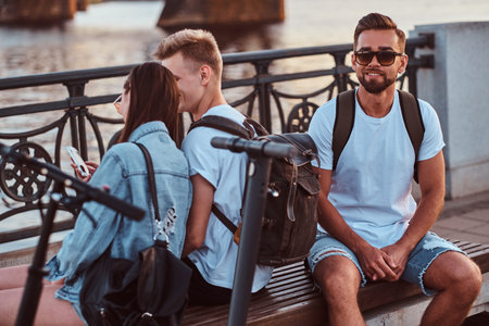 Three good happy friends are sitting on the bench at riverside. They have bagpacks and electronic...