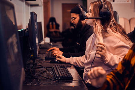 Focused girl get exited about videogame. She is wearing head set and glasses. another gamers at...