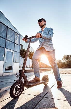 Low angle photo of smart elegant man in sunglasses and helmet which is driving his electrical scooter.の写真素材