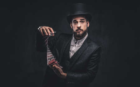 A Bearded magician in a black suit and top hat, showing trick with playing cards on a dark background.の写真素材