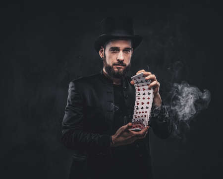 Magician in a black suit and top hat, showing trick with playing cards and magic smoke on a dark background.の写真素材
