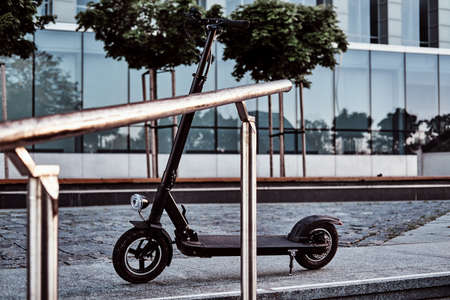 Nice black electrical scooter is parked near big glass building with offices.の写真素材