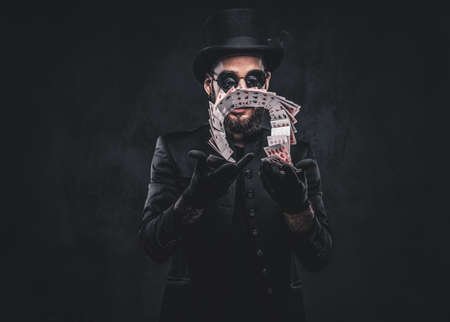 Magician in a black suit, sunglasses and top hat, showing trick with playing cards on a dark background.の写真素材